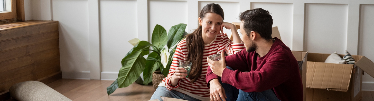 Casal sorrindo em ambiente doméstico, caixas de mudança ao fundo